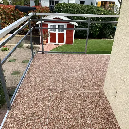 Balkon mit rot-weißen Steinteppich-Fliesen und Blick auf Gartenhaus im grünen Garten.